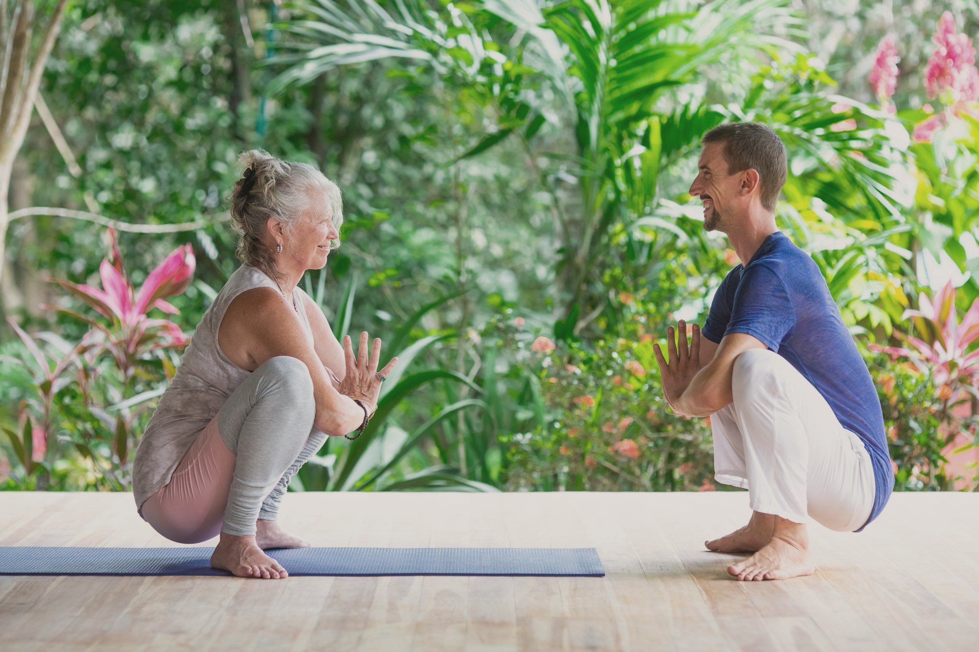 An older woman guest and a male instructor practice a guided yoga squat together in the jungle